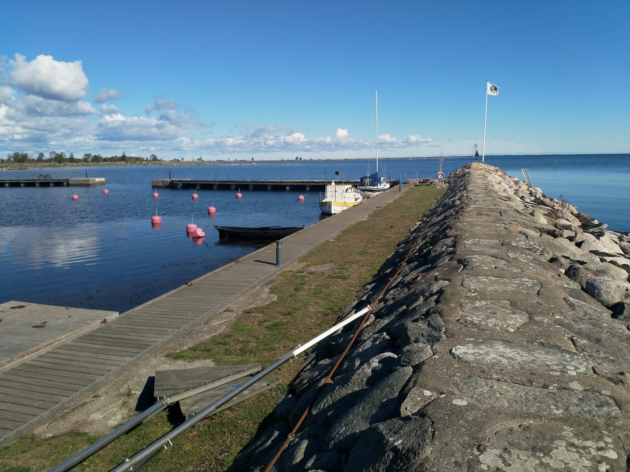 Steinmole führt ins Meer, links leere Stege, rote Bojen und vertäute Segelboote unter blauem Himmel.