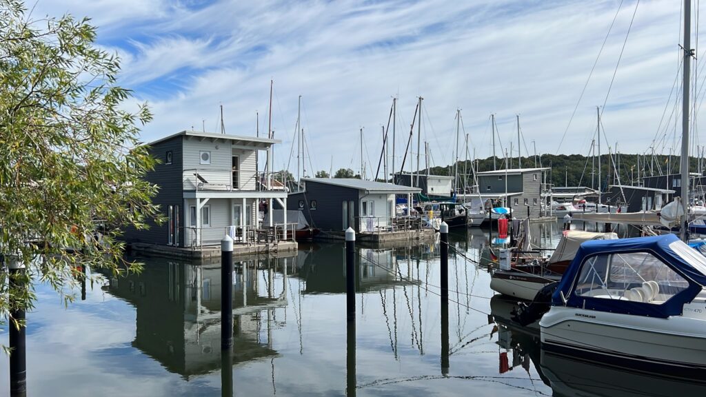 Schwimmende Häuser und Segelboote im Hafen, Spiegelungen tanzen auf glatter Wasseroberfläche unter blauem Himmel.