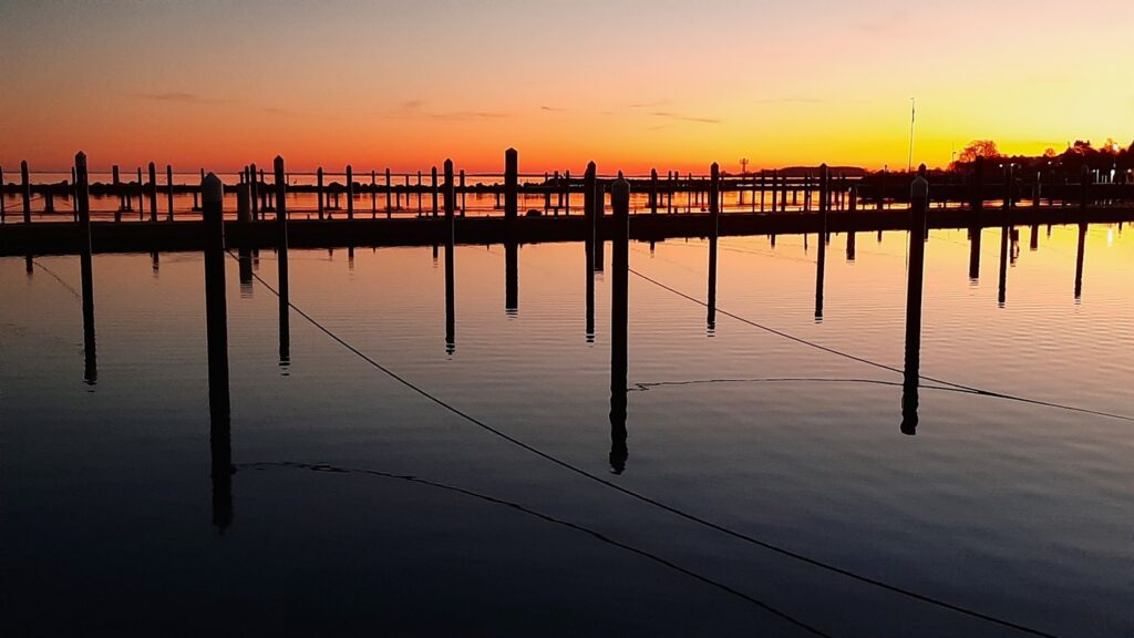 Stegpfosten spiegeln sich in ruhigem See, orange-violetter Sonnenuntergang am Horizont
