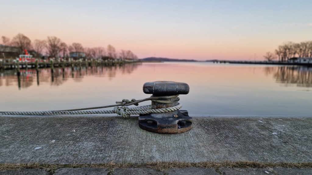 Festmachpoller mit vertäuter Leine am Kai, ruhiger Hafen spiegelt pastellfarbenen Sonnenuntergang.