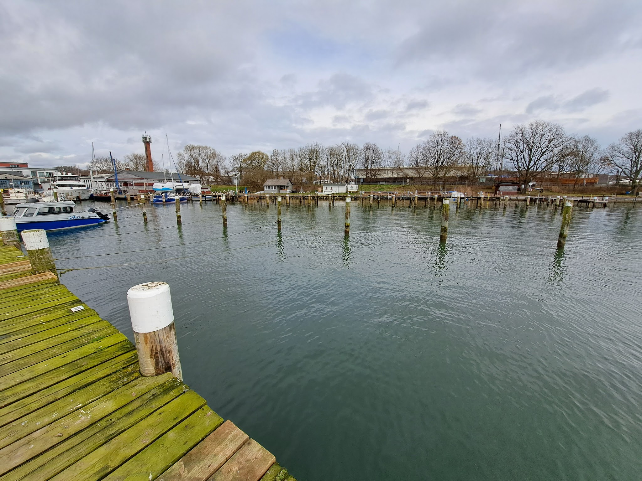 Leerer kleiner Hafen mit Holzsteg, vereinzelt angelegten Booten und Pfählen im ruhigen Wasser unter grauem Himmel.
