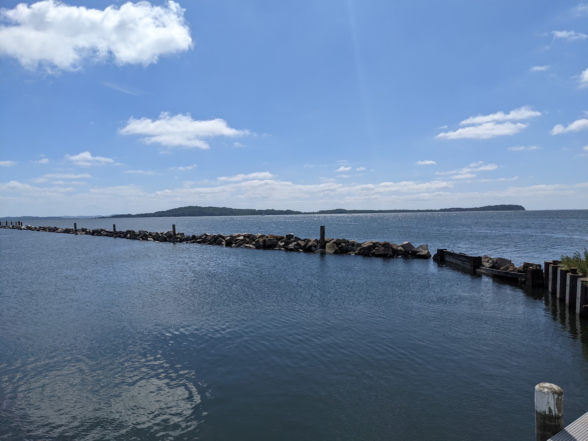 Steinmole ragt ins ruhige Wasser vor bewaldeter Insel unter blauem Himmel mit wenigen Wolken