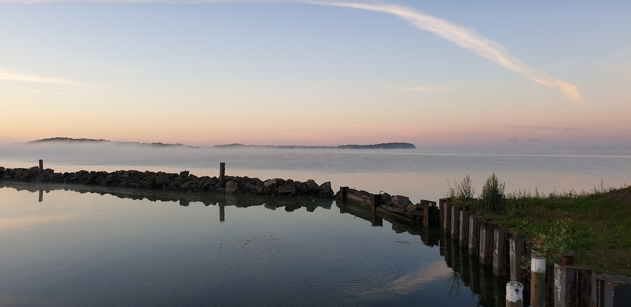 Ruhiger Sonnenaufgang an der Küste: Felsenmole, spiegelglattes Wasser mit Nebel, ferne Insel am Horizont.