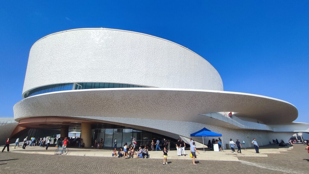 Weißes modernes, spiralartiges Gebäude mit breiter Rampe; Besucher stehen und sitzen davor unter blauem Himmel