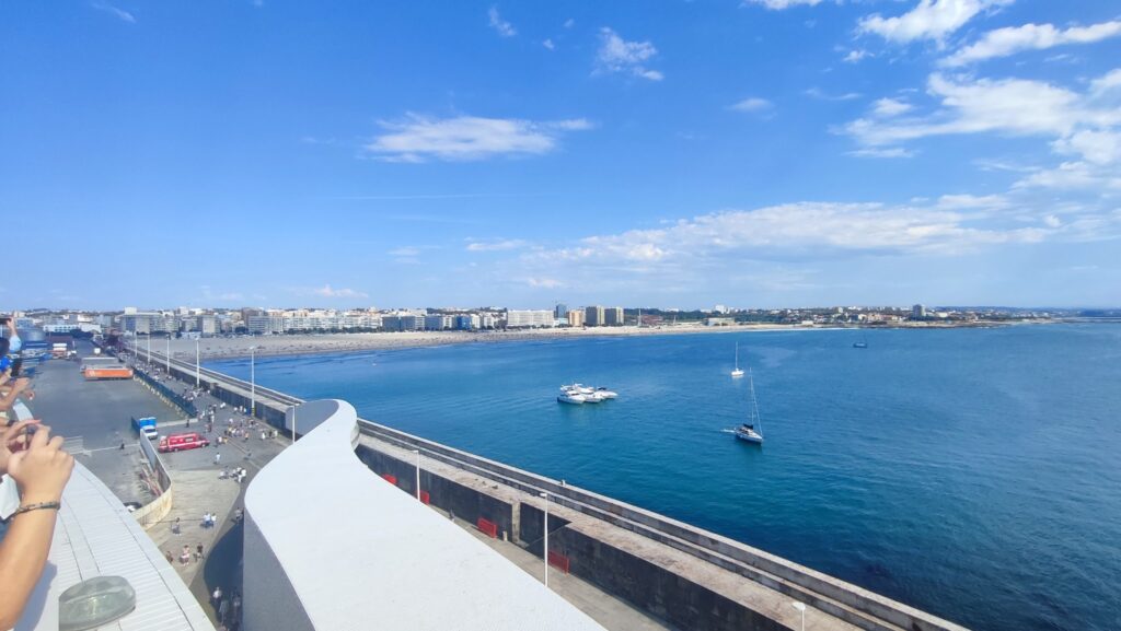 Blick von Plattform auf Küstenstadt, langen Strand und Hafen; ruhige blaue See mit drei Segelbooten unter klarem Himmel