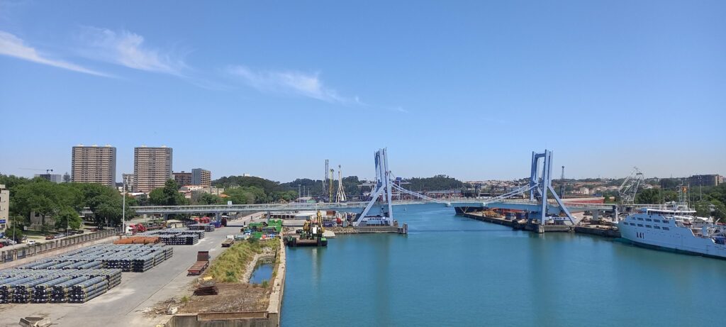 Hafen mit blauem Wasser, Klappbrücke, Frachtschiffen und Stapeln von Stahlrohren, Hochhäuser und Himmel im Hintergrund.
