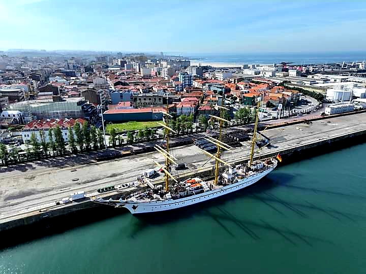Großes weißes Segelschulschiff mit drei gelb lackierten Masten liegt an Pier vor dicht bebauter Küstenstadt.