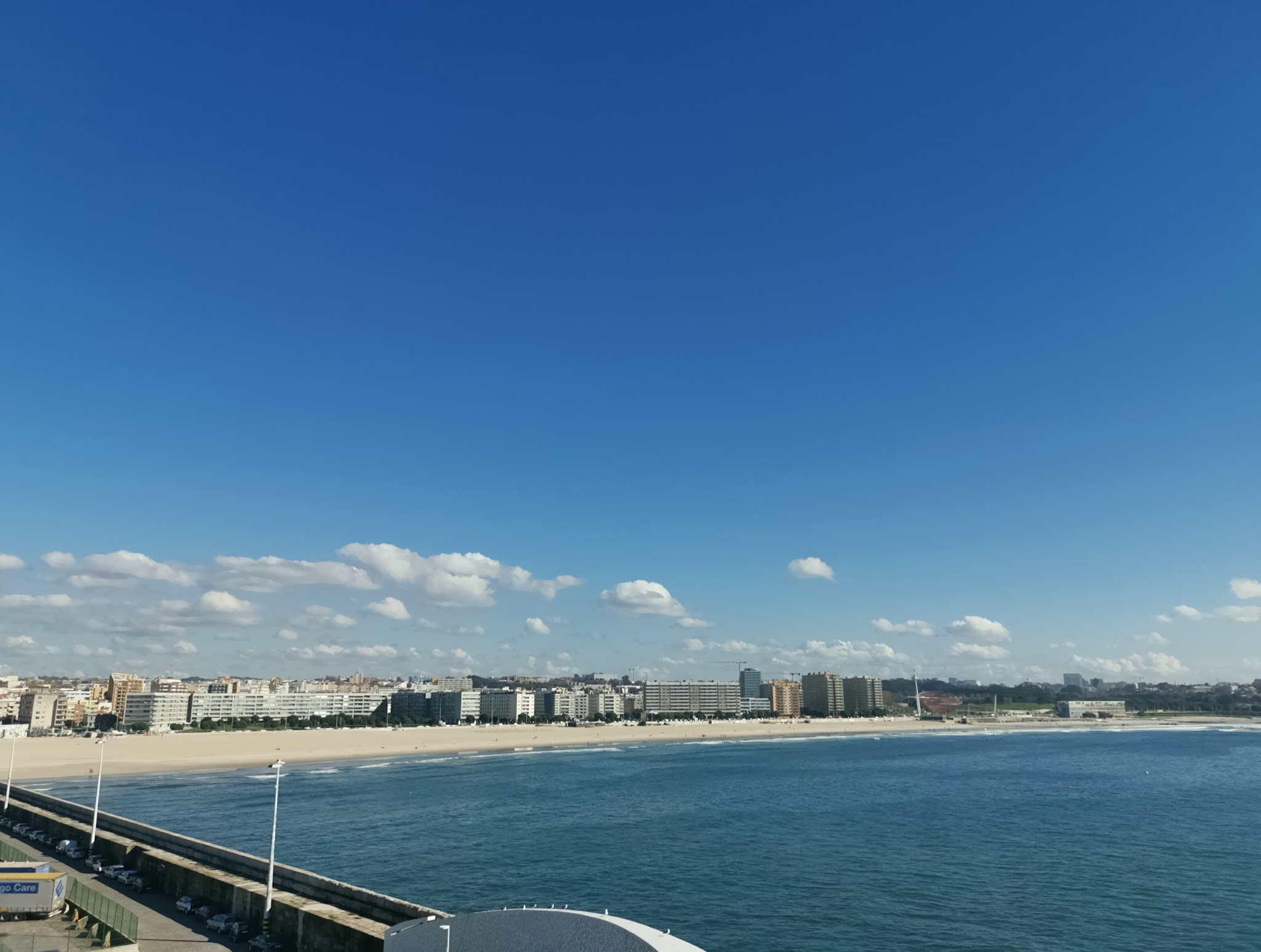 Weite Sandstrand mit Stadtpromenade, ruhiges blaues Meer, klarer Himmel mit wenigen Wolken.