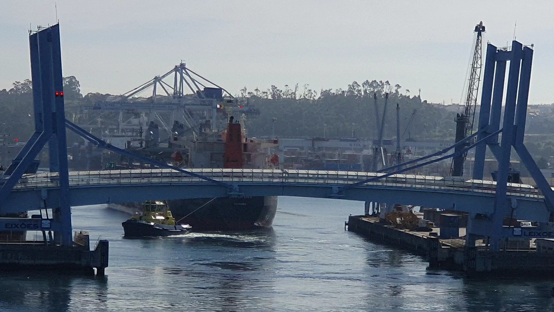 Gelber Schlepper zieht Frachtschiff unter blauer Hubbrücke im Hafen, Containerkräne im Hintergrund