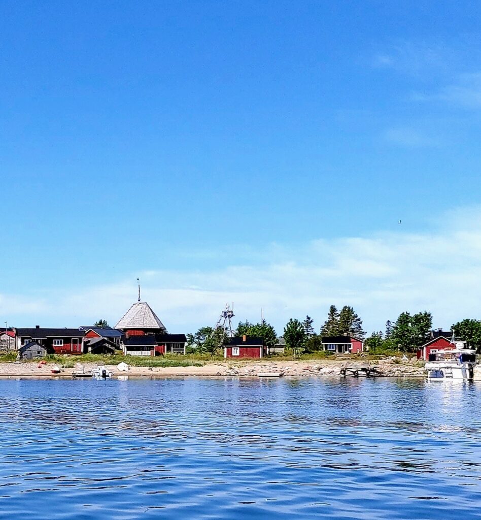 Rote Holzhäuser und ein Bootssteg an ruhiger Küstenlinie unter klarem Himmel, reflektiert im blauen Wasser
