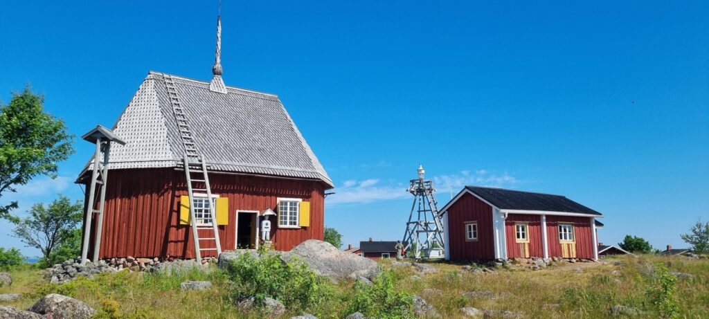 Kleine rote Holzhäuser mit Leiter, daneben schlanker Holzbaken-Leuchtturm auf felsiger, grasiger Küstenlandschaft.