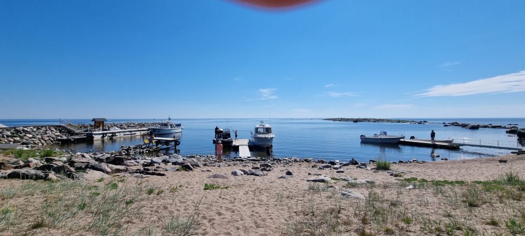 Kleiner Hafen: drei Motorboote an Stegen, ruhiges blaues Meer, sandiger Strand mit Felsen, klarer Himmel.