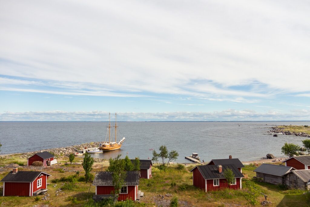 Holzsegler am Steg vor roten Fischerhütten an ruhiger Ostsee unter weitgezogenem Wolkenhimmel.