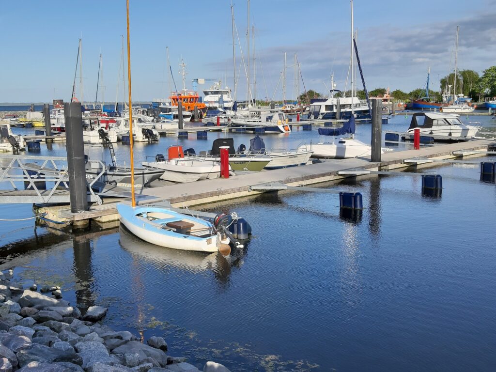 Sonniger Yachthafen: kleines weiß-blaues Ruderboot vorn, dahinter Reihen festgemachter Motor- und Segelboote.