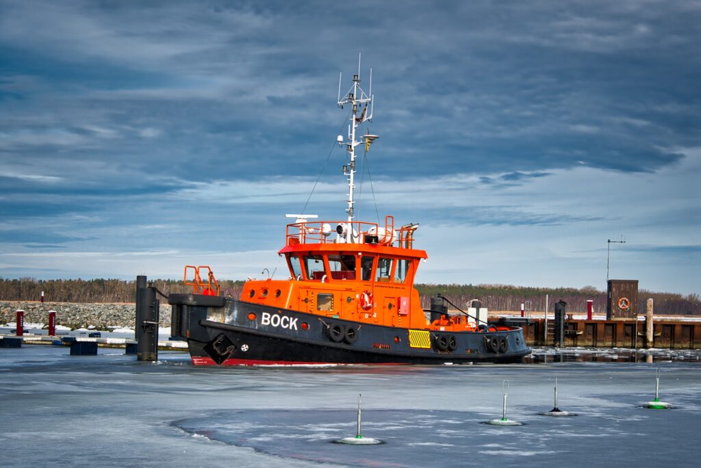 Orange Schlepper „BOCK“ liegt im teilweise zugefrorenen Hafen vor winterlicher Kulisse unter bewölktem Himmel.