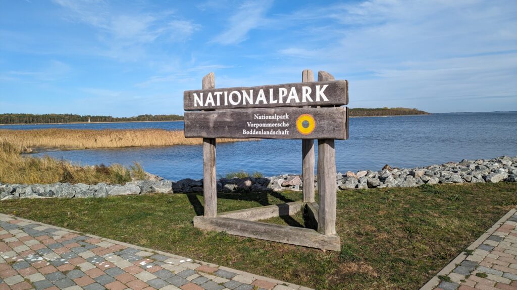 Holzschild „Nationalpark Vorpommersche Boddenlandschaft“ am Boddenufer mit Schilf, Steinen und weitem Himmel