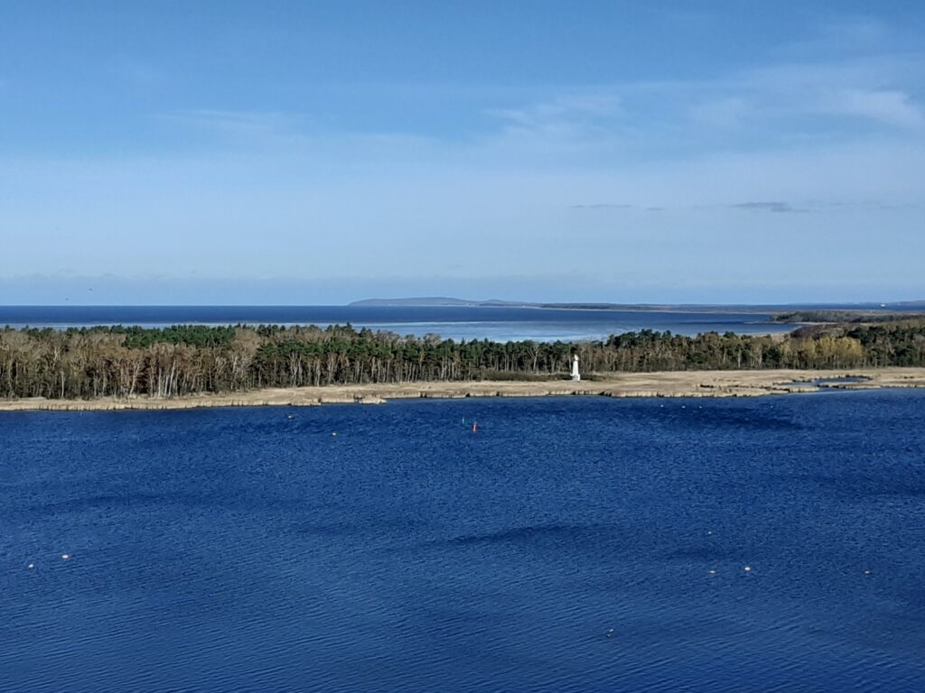 Tiefer blauer See, vorgelagerte Waldinsel mit kleinem weißem Leuchtturm, dahinter offenes Meer unter blauem Himmel.