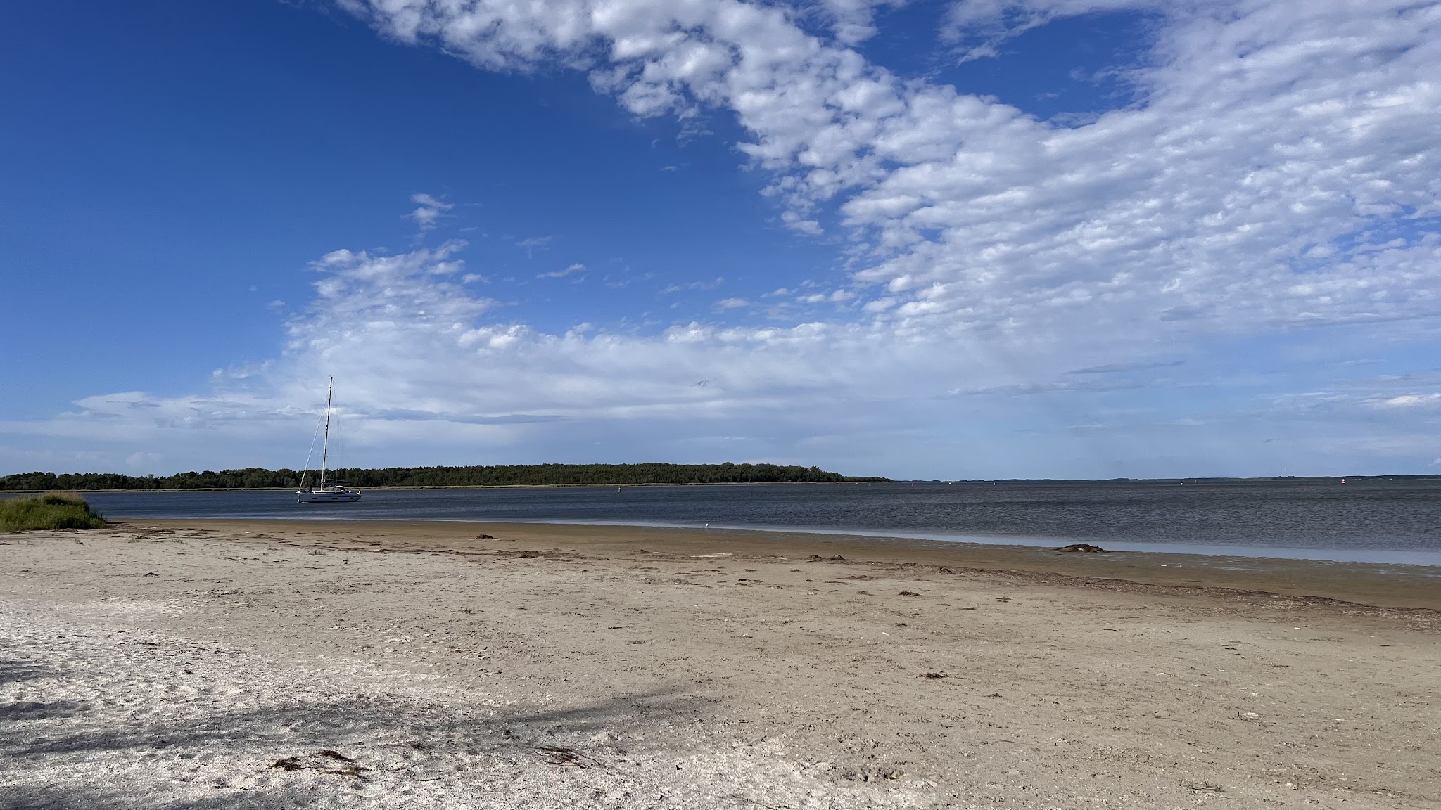 Weitläufiger Sandstrand, stilles Meer, einzelnes Segelboot vor bewaldeter Küstenlinie unter locker bewölktem Himmel