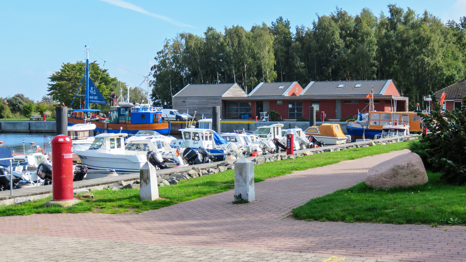 Marina mit Reihe kleiner Motorboote und buntem Fischkutter; Gehweg vorn, dahinter Bootshäuser und Bäume.
