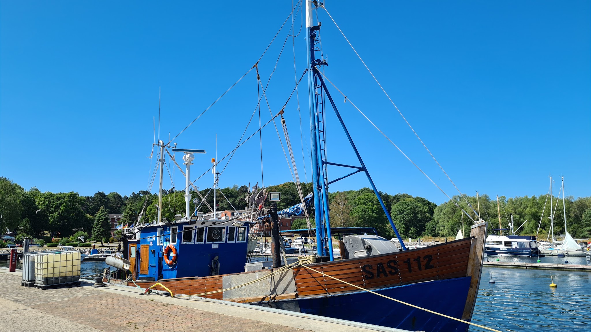 Fischkutter „SAS 112“ liegt bei Sonnenschein am Kai; ruhiger Hafen, Segelboote, grüne Uferbäume