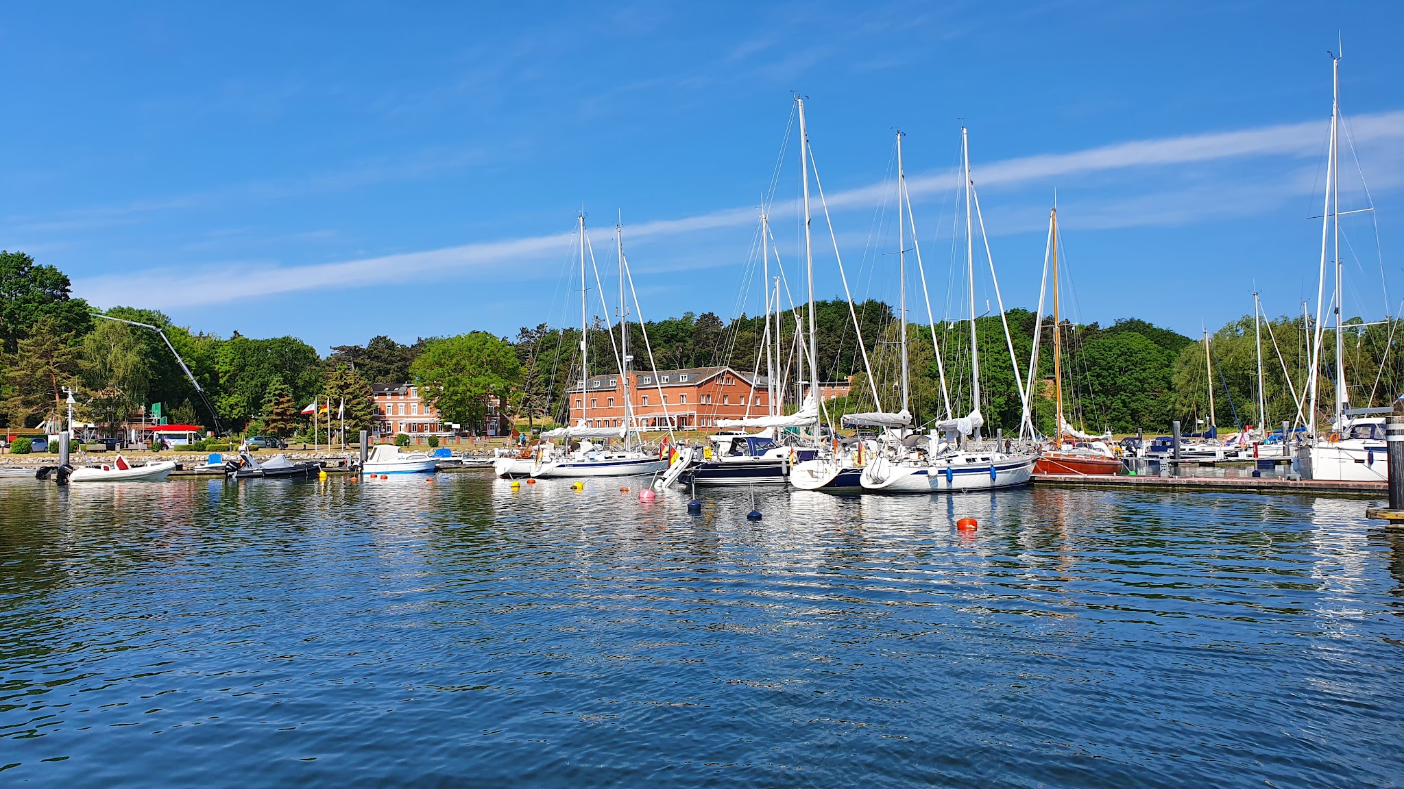 Segelboote liegen im ruhigen Hafen vor roten Backsteingebäuden und grünen Bäumen unter klarem, blauem Himmel.