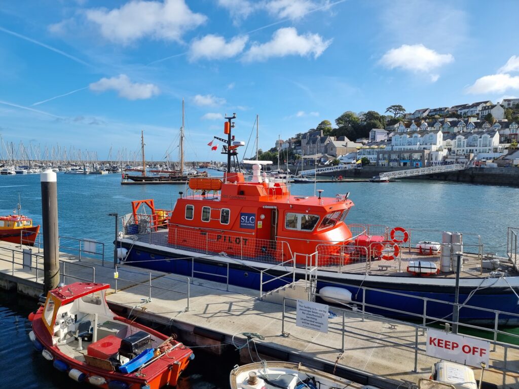 Orangefarbenes Lotsenboot am Kai; kleinere Boote, Mastwald der Marina und Küstenstädtchen im Hintergrund.