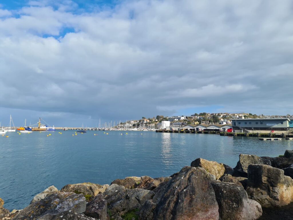 Hafen mit Segelbooten, Felsen im Vordergrund, Küstenstädtchen und bewölkter Himmel im Hintergrund