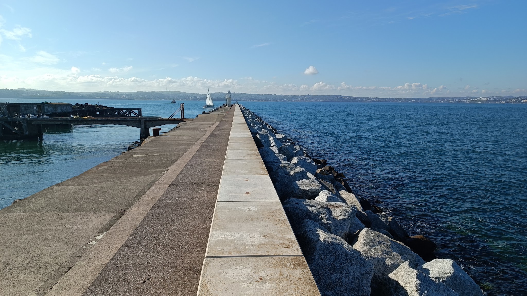 Langer Pier mit Steinschutz führt zum weißen Leuchtturm; Segelboot daneben, ruhiges blauschimmerndes Meer.