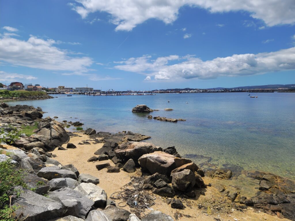 Steinige Küste mit klarem, blauem Meer; im Hintergrund Hafen und Boote unter locker bewölktem Himmel