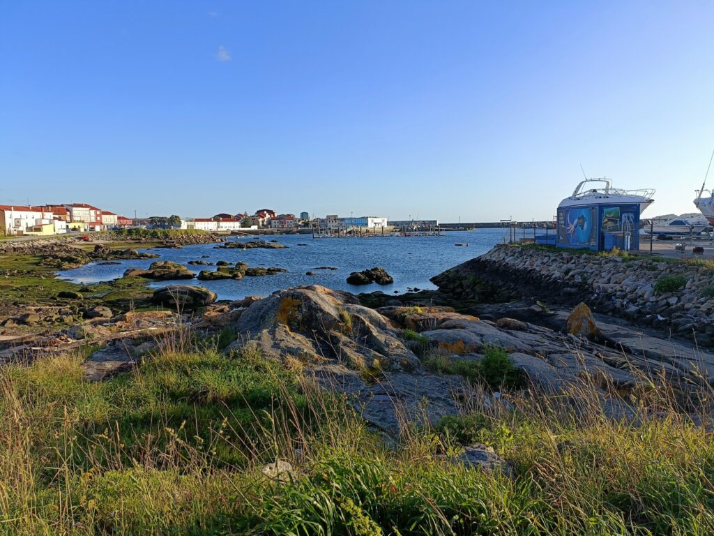 Felsige Küste mit Gras, ruhiger Hafen, bunte Häuserreihe links, Bootswerft mit Yacht rechts unter blauem Himmel
