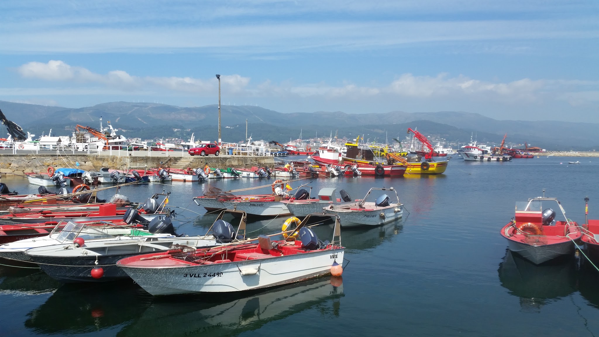 Kleiner Hafen: mehrere rot-weiße Fischerboote liegen im ruhigen Wasser vor einer Mole, Berge im Hintergrund.