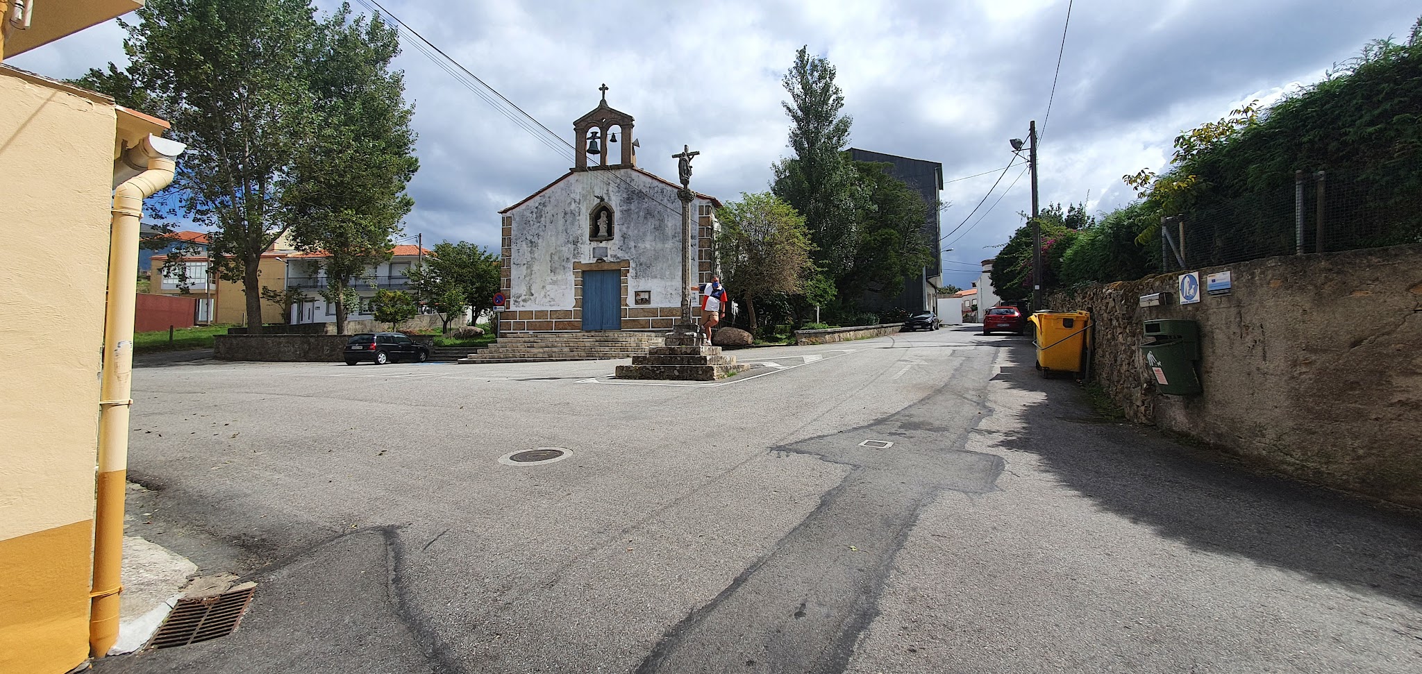 Kleine Dorfkapelle mit Glockengiebel; davor Kreuz und Stufen, ruhige Straße, Person mit buntem Regenschirm.
