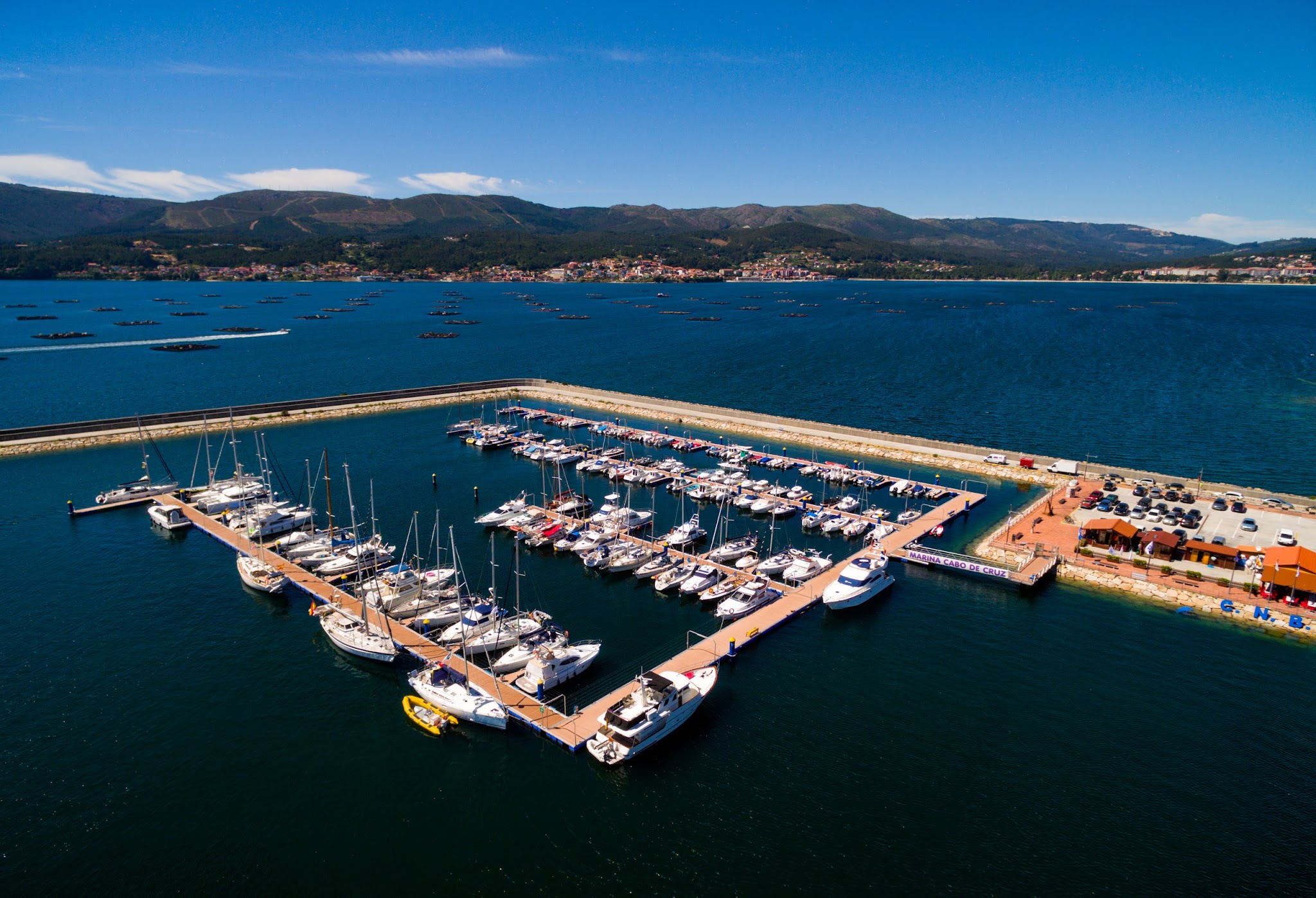 Luftaufnahme einer Marina: Docks mit zahlreichen Segelbooten und Yachten, umgeben von blauem Meer, Berge am Horizont.