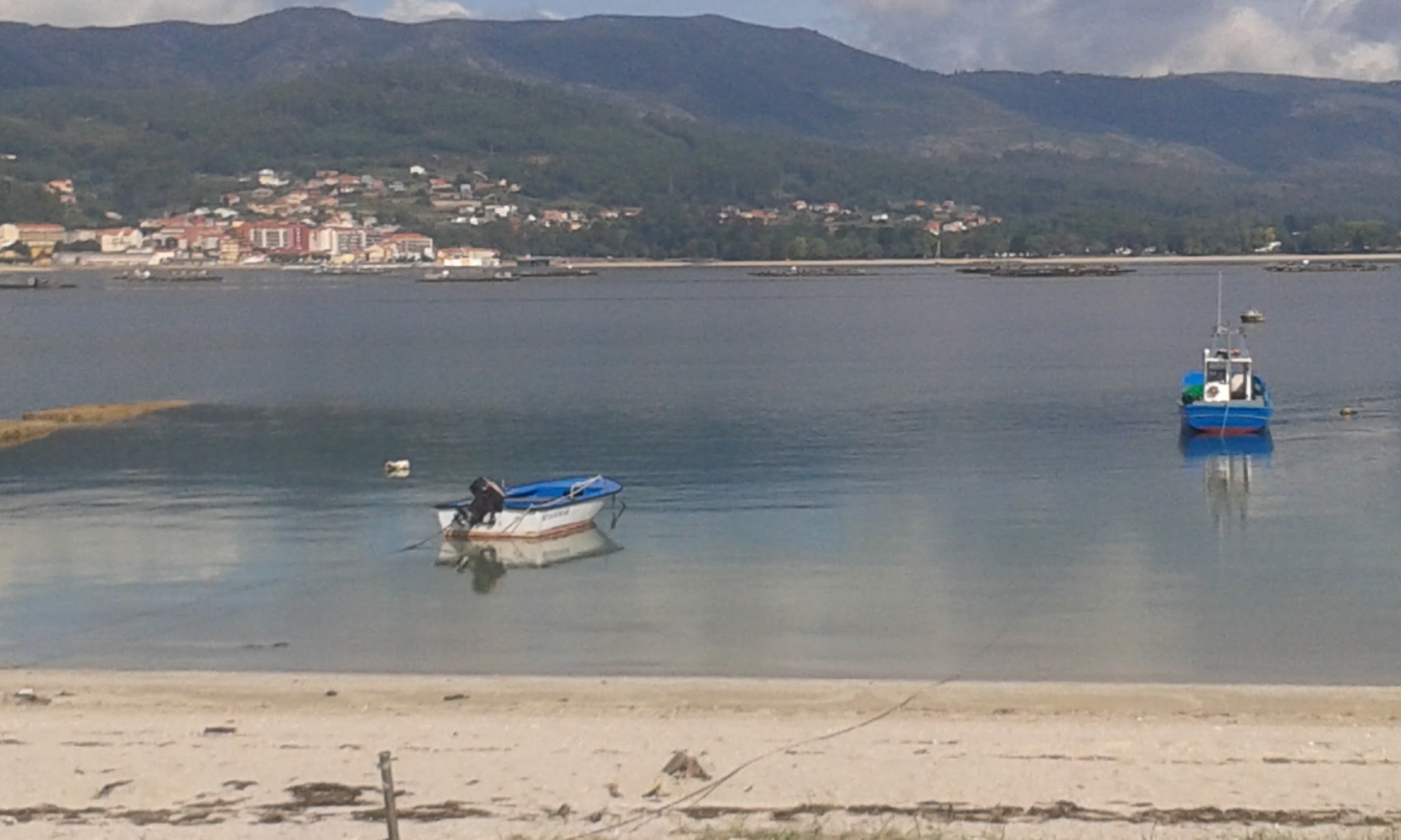 Zwei Boote liegen still in einer ruhigen Bucht; vorn Sandstrand, dahinter Küstenstadt vor grünen Bergen.