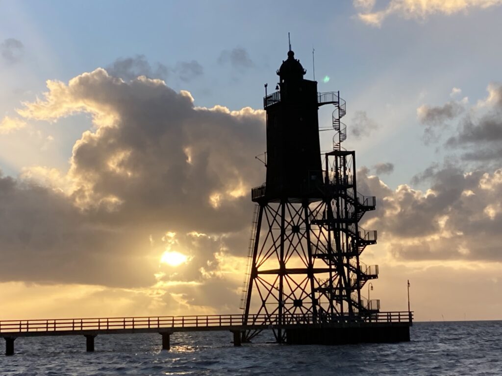Stählerner Leuchtturm auf Stelzen im Meer, Sonnenuntergang durch Wolken, Steg führt zum Turm.