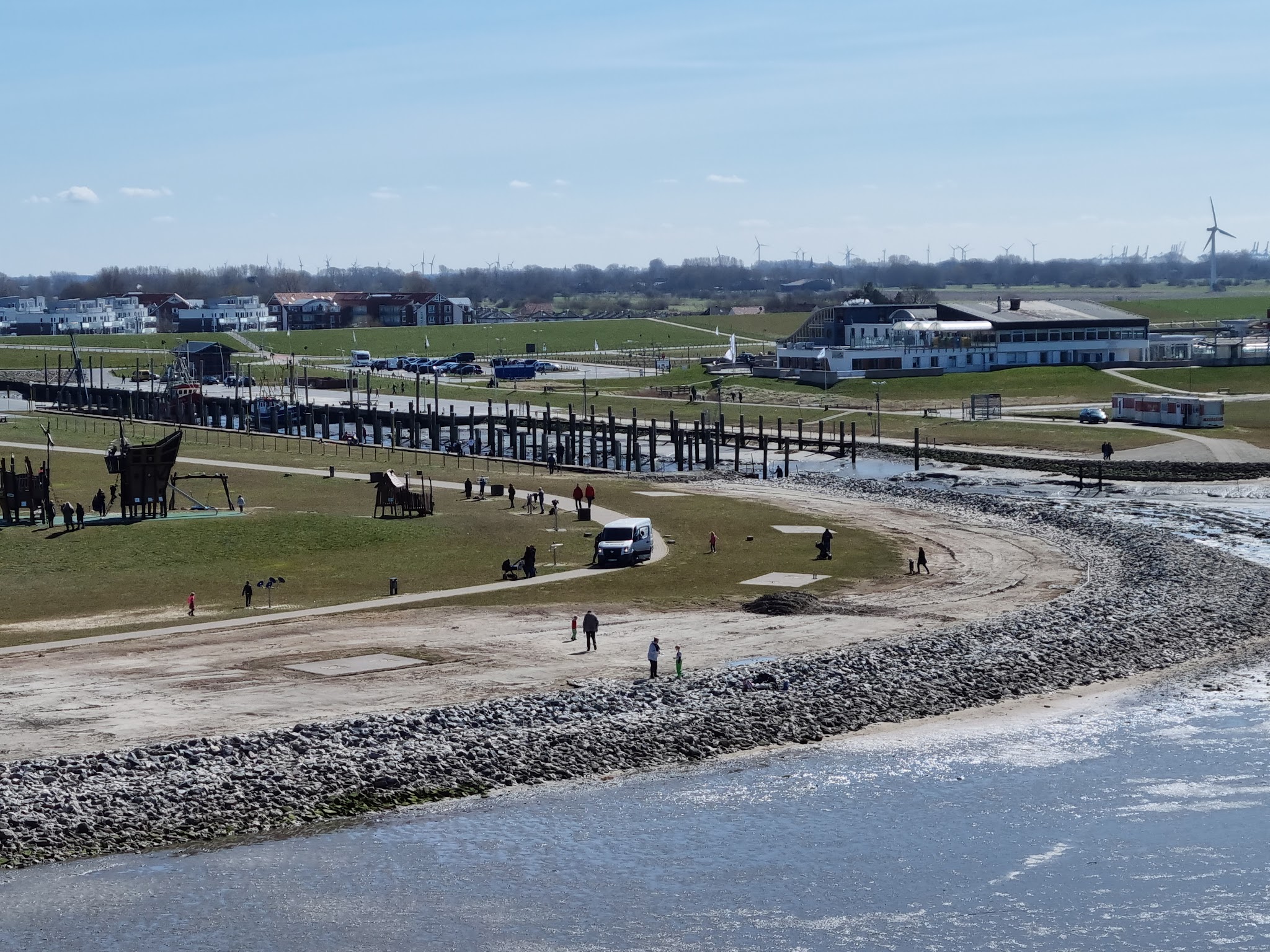 Küstenspielplatz mit Holzschiff, Spaziergänger am Strand, Hafensteg, im Hintergrund Deich, Häuser und Windräder.