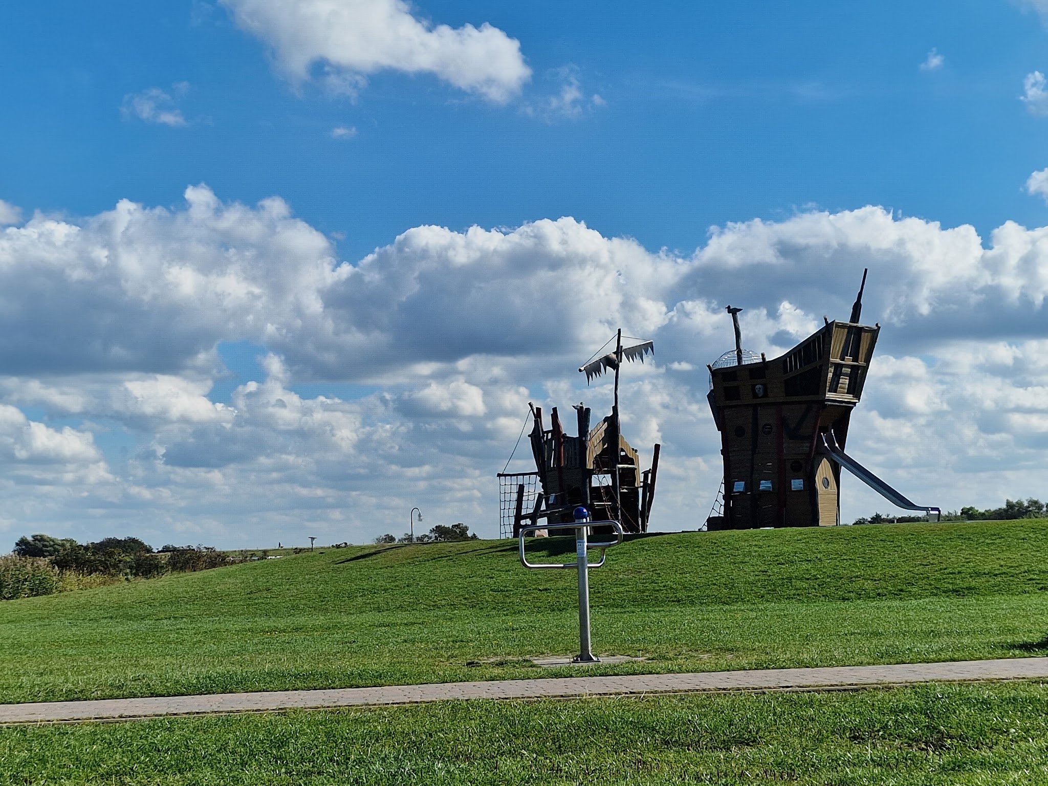 Holz-Piratenschiff-Spielplatz auf sanftem grünen Hügel unter blauem Himmel mit lockeren Wolken.