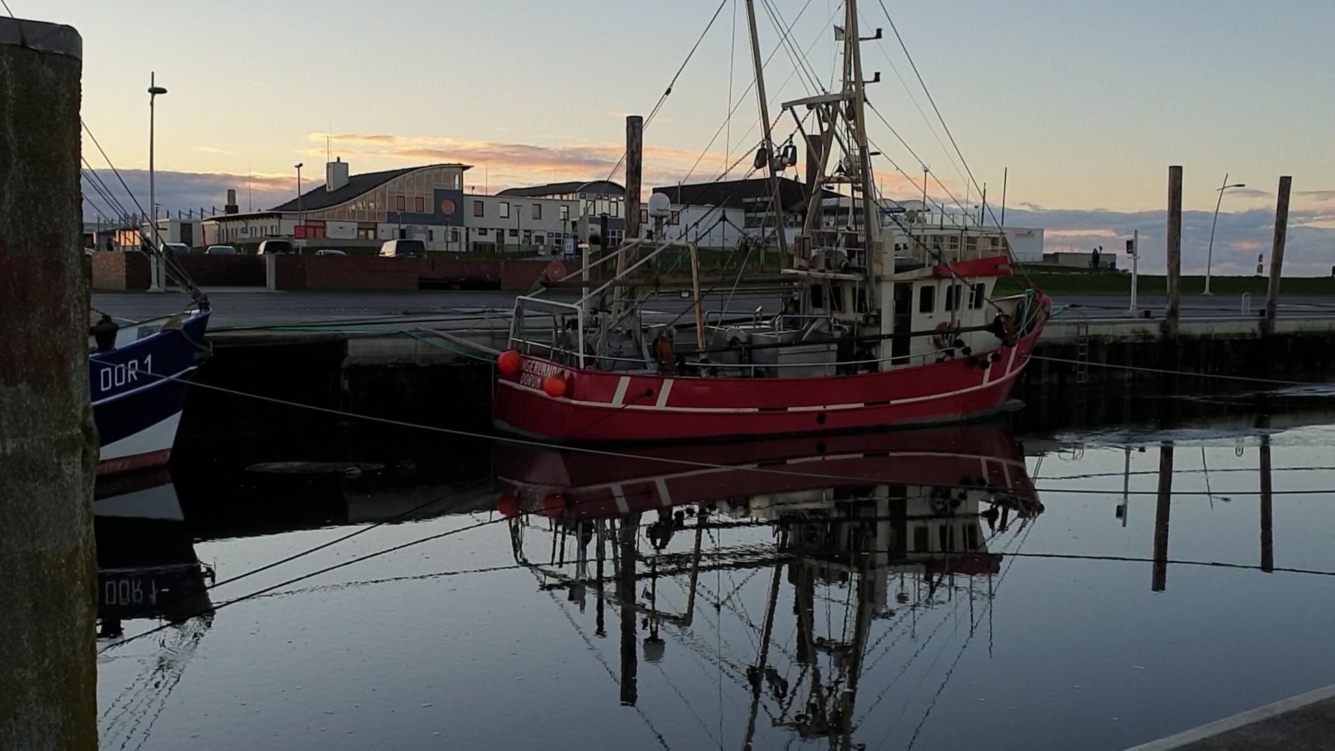 Rotes Fischerboot liegt bei ruhiger Abenddämmerung im Hafen, Mast und Rumpf spiegeln sich klar im stillen Wasser