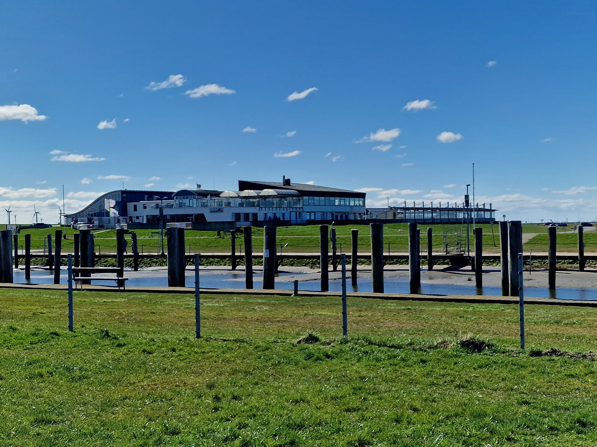 Grünes Ufer mit leeren Hafenpfählen, modernes Küstengebäude und Windräder unter blauem Himmel.
