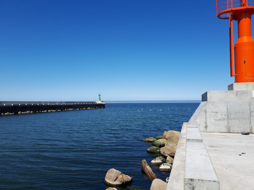 Hafenmole mit grünem Leuchtturm links; im Vordergrund rechts massiver orangefarbener Turm, ruhige See, blauer Himmel