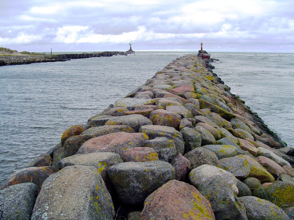 Langer Steinwellenbrecher führt durchs graue Meer zu kleinem Leuchtturm unter bewölktem Himmel.