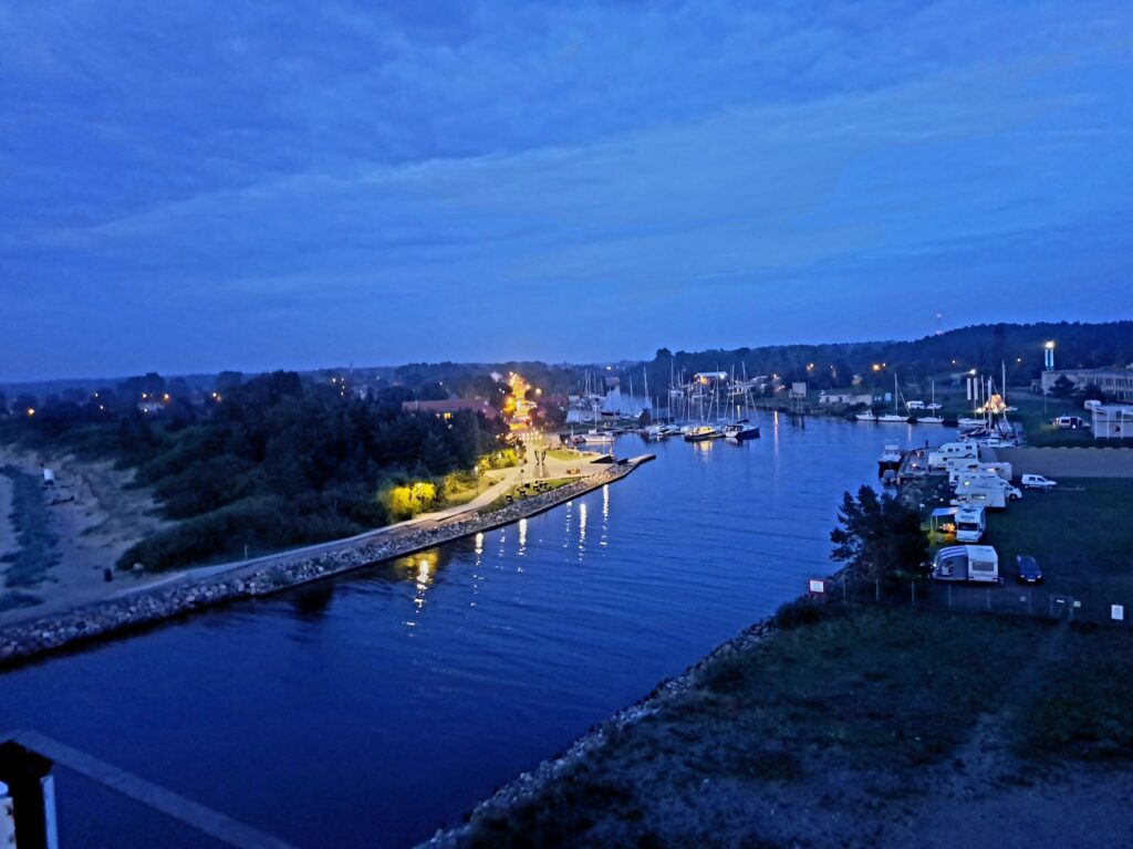 Blaues Abendpanorama: beleuchteter Hafenkanal, Segelboote, Promenade, rechts parkende Wohnmobile am Ufer