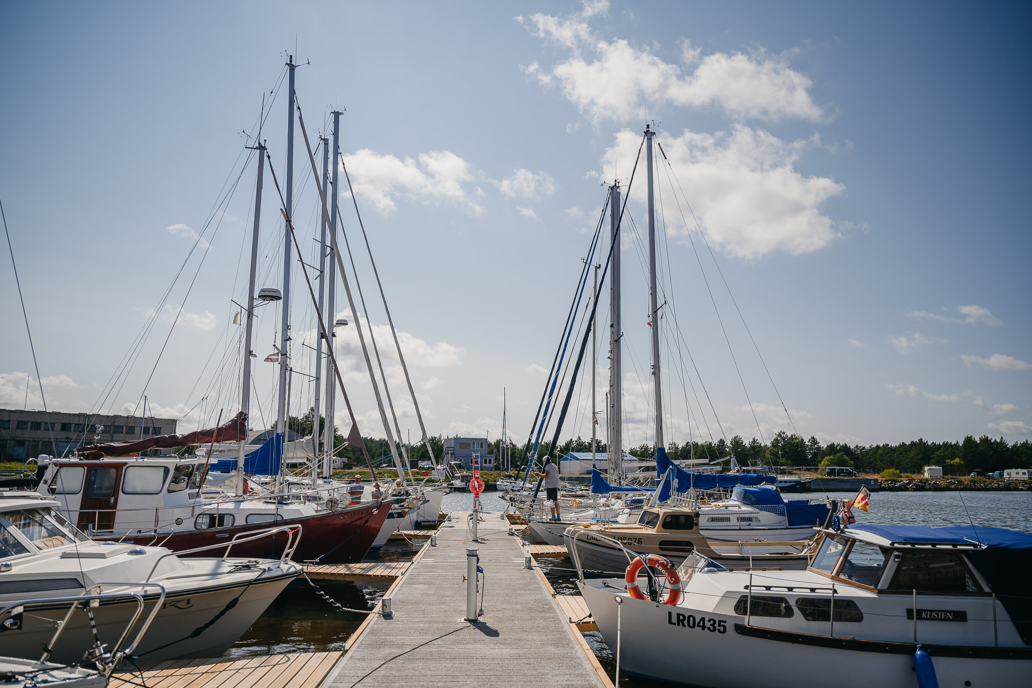 Steg mit beidseitig vertäuten Segelbooten im sonnigen Hafen; hohe Masten ragen in blauen Himmel.