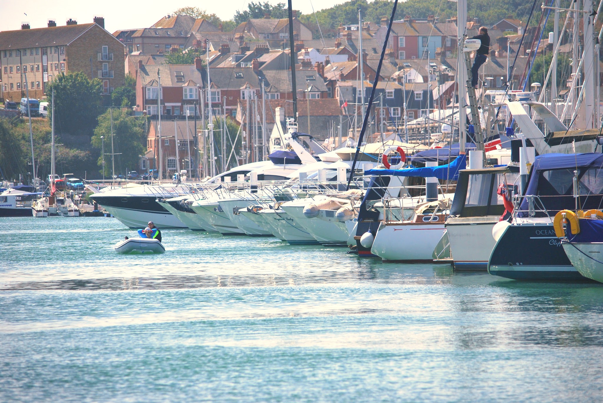 Mehrere Yachten im Hafen; vorne fährt ein Schlauchboot mit zwei Personen über ruhiges Wasser vor Stadtkulisse.