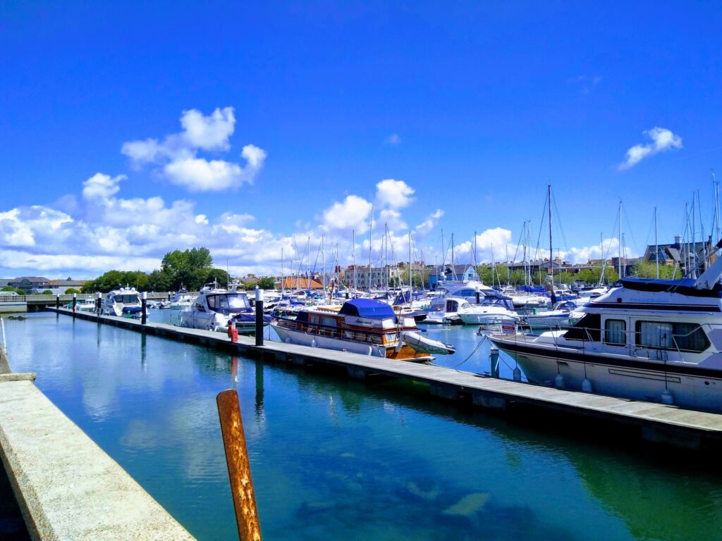 Sonniger Jachthafen; zahlreiche Segelboote und Yachten liegen ruhig am Steg, blauer Himmel mit weißen Wolken.