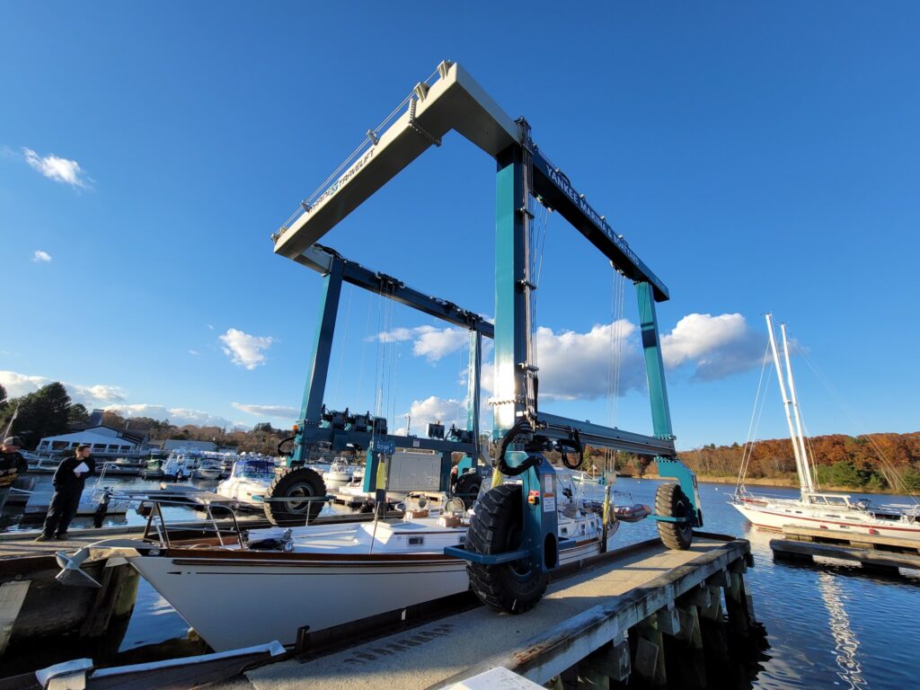 Mobiler blauer Bootskran hebt Segelboot am Dock; Yachthafen mit weiteren Booten unter klarem Himmel.