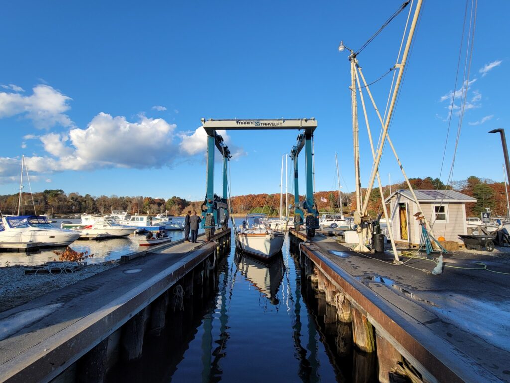 Segelboot hängt in blauem Travellift zwischen zwei Stegen im Jachthafen; sonniger Herbst, Boote im Hintergrund