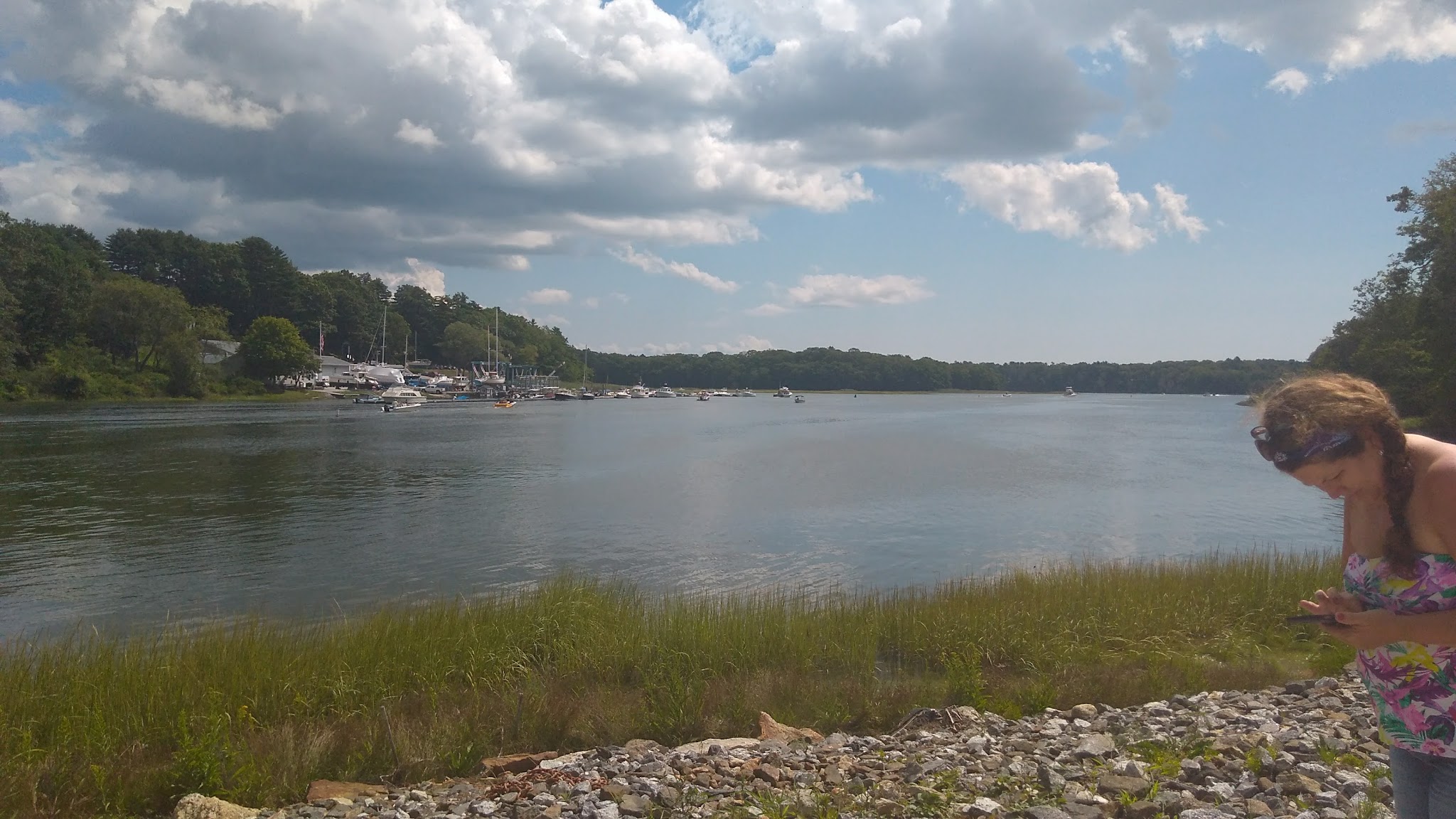 Ruhiger Hafenarm mit Booten unter bewölktem Himmel; vorne rechts Frau tippt am Ufer auf ihr Smartphone.