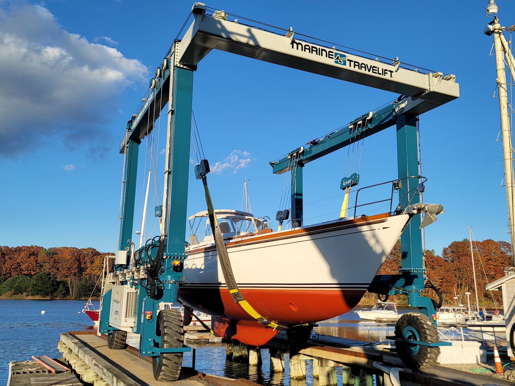 Bootslift hebt weißes Segelboot über Steg, blauer Himmel und herbstliche Bäume im Hintergrund