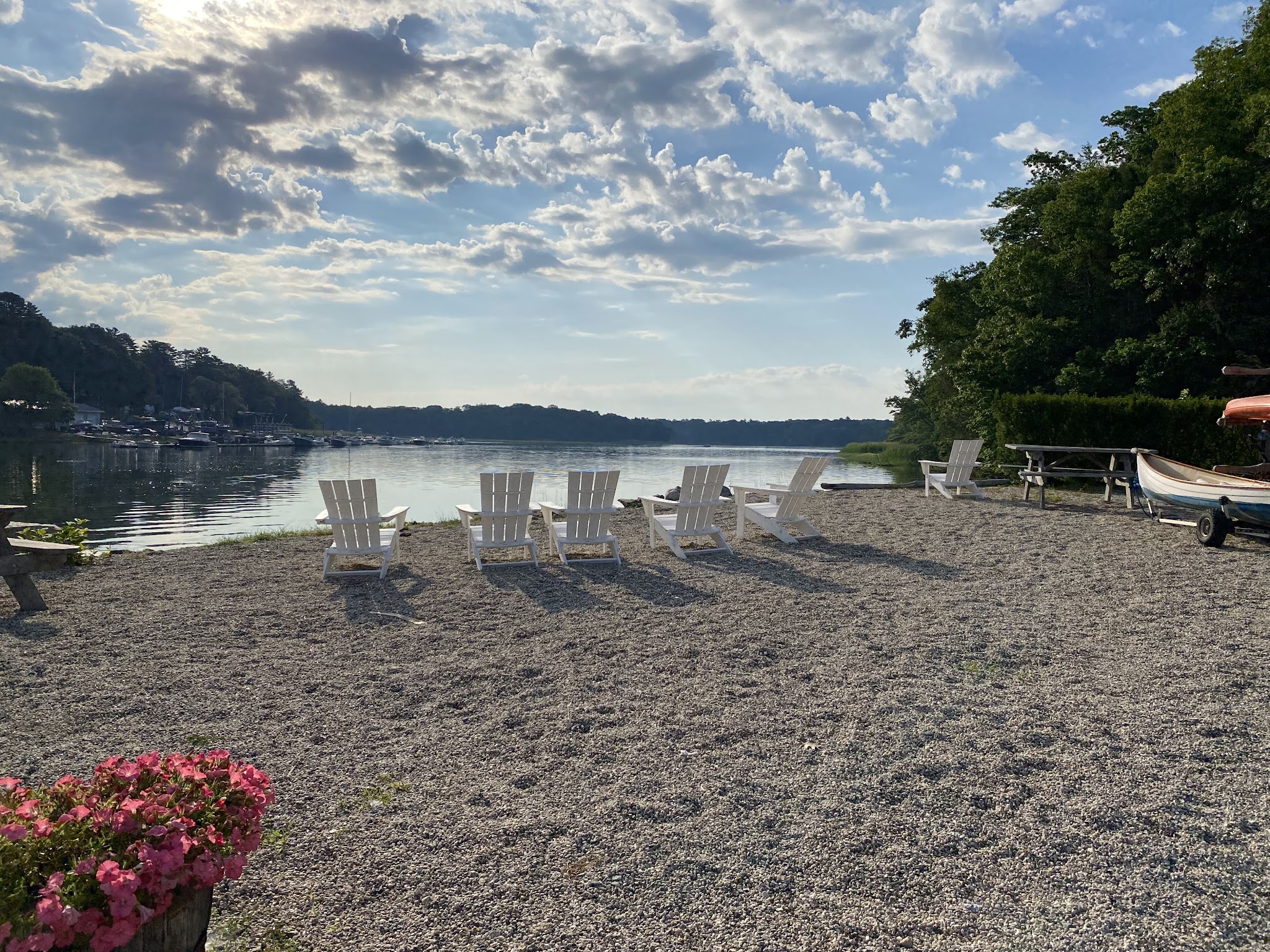 Mehrere weiße Liegestühle auf Kiesufer blicken über ruhigen See; links Boote, rechts Bäume, sommerliche Wolken.